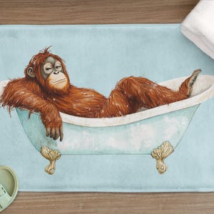Peut inclure: Un tapis de bain bleu clair avec une illustration de dessin animé d'un orang-outan prenant un bain dans une baignoire blanche avec des pieds dorés. L'orang-outan est allongé sur le dos, les bras reposant sur le bord de la baignoire.