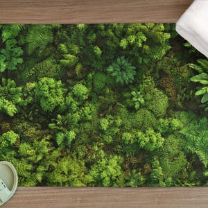 Può includere: Un tappetino da bagno rettangolare con un fitto motivo botanico verde, che ricorda una foresta lussureggiante. Il tappetino è posto su una superficie di legno, con sandali verde chiaro e un asciugamano bianco.