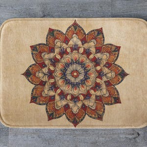 May include: A rectangular beige bath mat with a detailed mandala design in red, orange, blue, and cream. The floral pattern is centered on the mat, creating a symmetrical design. The mat has rounded corners.