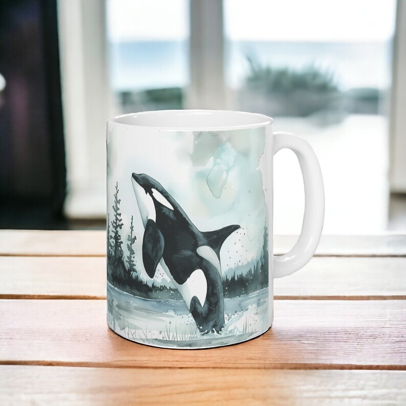 May include: White ceramic mug with a watercolor illustration of an orca jumping out of the water. The orca is black and white with a splash of water behind it. The background is a light blue watercolor wash with a few trees in the background.