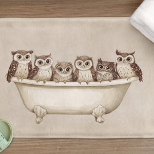 Peut inclure: Un tapis de bain beige avec une illustration à l'aquarelle de six hiboux dans une baignoire vintage. Les hiboux ont des plumes marron et blanches et de grands yeux. Une serviette blanche repose sur le tapis, et des sandales vertes sont visibles.