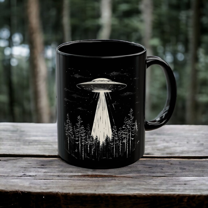 May include: Black ceramic coffee mug featuring a white graphic of a UFO flying over a forest at night.