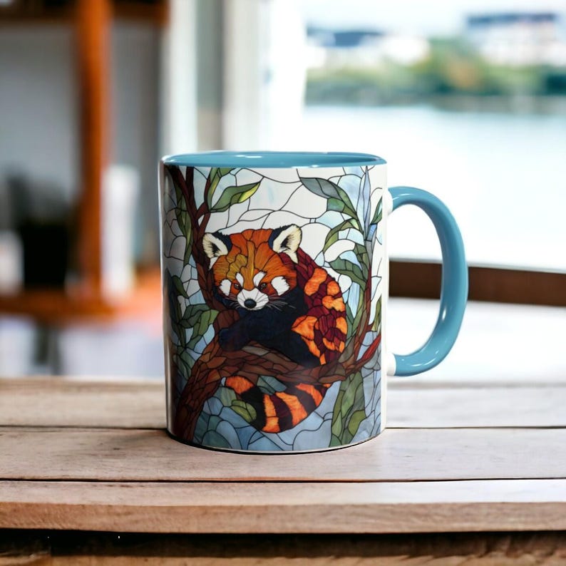 Puede incluir: Una taza de cer&aacute;mica con un dise&ntilde;o de vidriera que representa un panda rojo posado en la rama de un &aacute;rbol. La taza tiene un asa y un interior azul claro, y est&aacute; sobre una superficie de madera.
