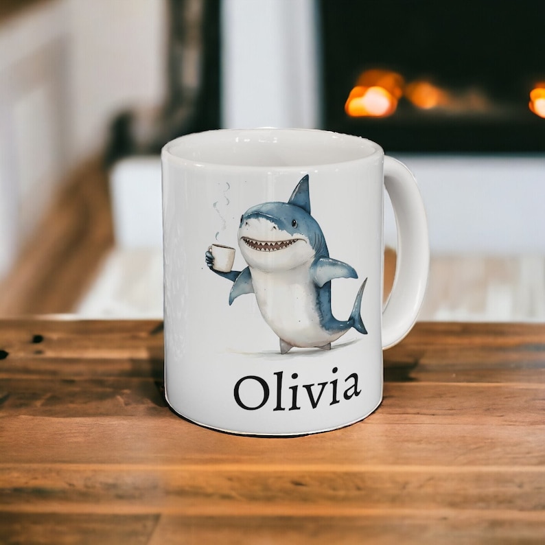 May include: White ceramic mug with a cartoon illustration of a shark holding a cup of coffee. The shark is blue and gray and has a toothy grin. The mug has the name "Olivia" printed on the bottom.