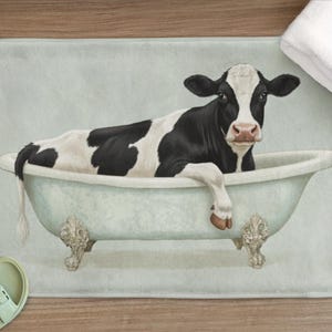 May include: A novelty bath mat depicting a black and white cow in a vintage bathtub. The mat has a light blue background, with the cow as the central image. The tub has decorative claw feet. A white towel and sandals are also visible.