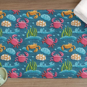 May include: A rectangular bath mat featuring an embroidered pattern of crabs, seaweed, and shells on a teal background. The crabs are pink and orange. A white towel is on the right. Sandals are on the left.
