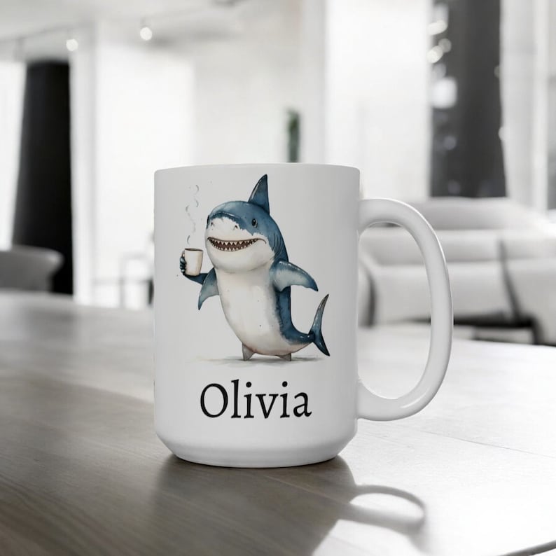 May include: White ceramic mug with a cartoon illustration of a shark holding a cup of coffee. The mug has the name "Olivia" printed on the bottom.