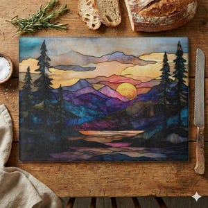 May include: A rectangular glass cutting board with a sunset landscape design. The artwork features a yellow sun, purple mountains, and dark green trees. The board is surrounded by a wooden table, bread, a knife, and herbs.
