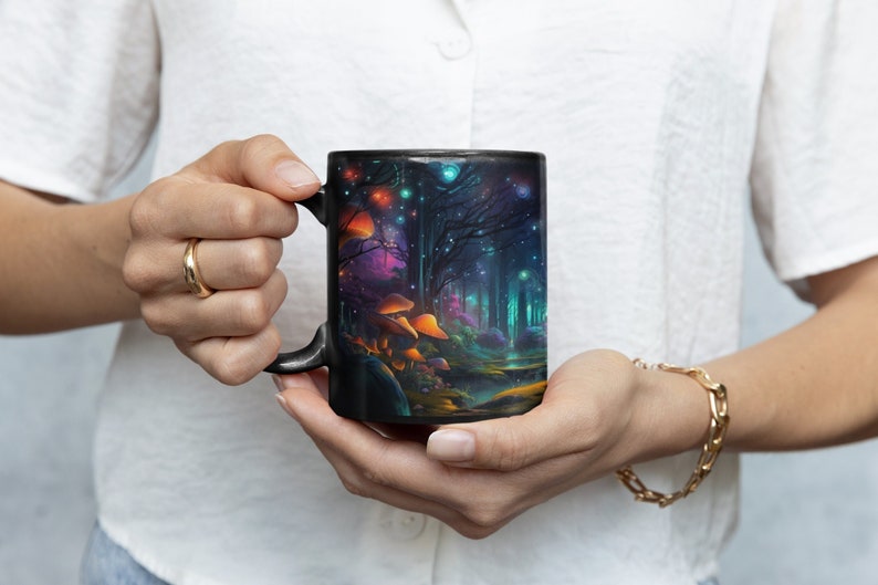 K&ouml;nnte beinhalten: Schwarze Keramiktasse mit einer farbenfrohen Illustration einer magischen Waldszene mit leuchtenden Lichtern, Pilzen und B&auml;umen.