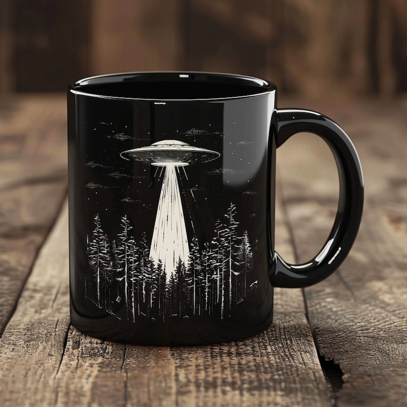May include: Black ceramic coffee mug featuring a white graphic of a UFO flying over a forest. The UFO is emitting a beam of light.