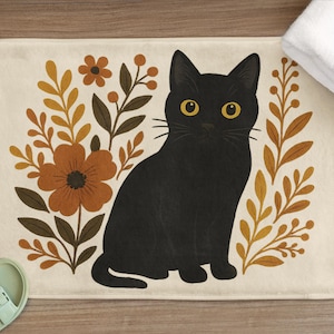 Puede incluir: Un gato negro con ojos amarillos se sienta en el centro de una alfombra de baño de color crema. El gato está rodeado de diseños florales estilizados en marrón y amarillo.