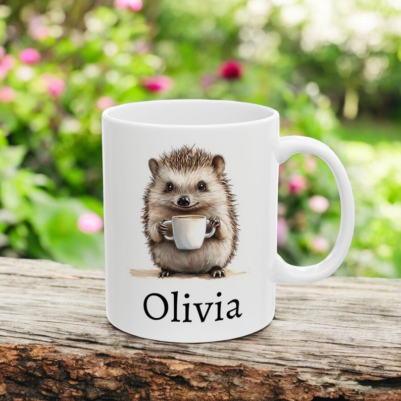 May include: White ceramic mug with a hedgehog illustration holding a cup of coffee. The mug has the name "Olivia" printed on the bottom.