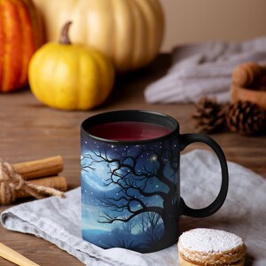 May include: A black coffee mug with a spooky forest scene printed on it. The mug is filled with a dark red liquid and is sitting on a gray linen napkin. There is a cookie with powdered sugar on top next to the mug.