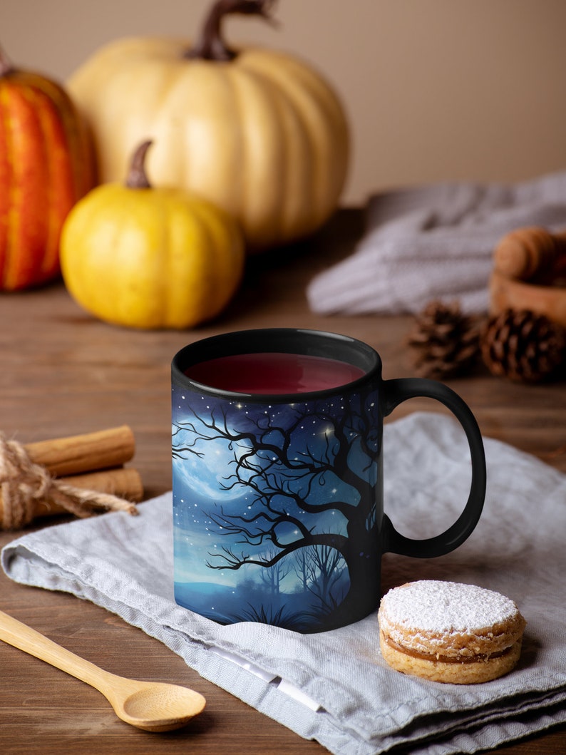 May include: A black coffee mug with a spooky forest scene printed on it. The mug is filled with a dark red liquid and is sitting on a gray linen napkin. There is a cookie with powdered sugar on top next to the mug.