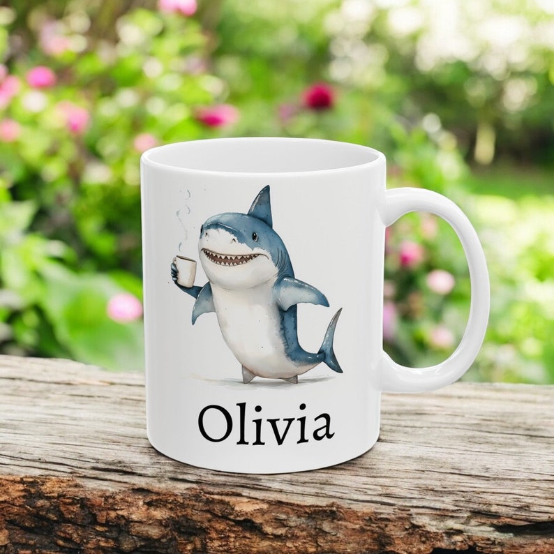 May include: White ceramic mug with a cartoon illustration of a blue shark holding a cup of coffee. The mug has the name "Olivia" printed on the bottom.