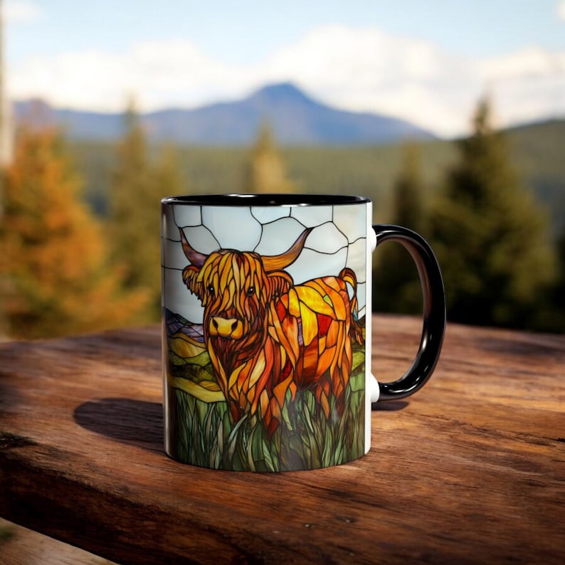 May include: A white ceramic mug with a black rim and handle. The mug features a stained glass design of a Highland cow with brown, orange, and yellow colors.