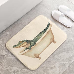Puede incluir: Una alfombra de baño beige con un cocodrilo verde en una bañera blanca. El cocodrilo está mirando hacia la izquierda. La alfombra de baño está sobre un suelo de baldosas de color gris claro.