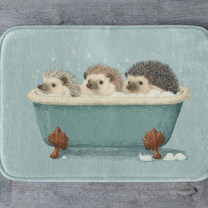 Puede incluir: Alfombra de baño rectangular con una ilustración caprichosa de tres erizos relajándose en una bañera turquesa llena de burbujas. La bañera tiene patas marrones ornamentadas. El fondo es de color turquesa claro.