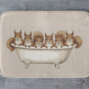 Könnte beinhalten: Eine rechteckige Badematte mit einer Aquarellillustration von sechs braunen Eichhörnchen in einer weißen Badewanne mit Löwenfüßen. Die Eichhörnchen schauen nach vorne, ihre Pfoten liegen am Wannenrand. Der Hintergrund ist hellbeige.