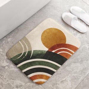 Può includere: Un tappetino da bagno geometrico con un design a sole e paesaggio nei toni del beige, verde, arancione e nero. Il tappetino è posizionato su un pavimento piastrellato grigio vicino a una vasca da bagno bianca e un paio di pantofole bianche.