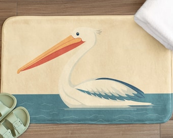 May include: A bath mat featuring a cartoon illustration of a pelican swimming in blue water. The pelican is white with a long orange beak and blue accents on its wings. The background is a light beige.