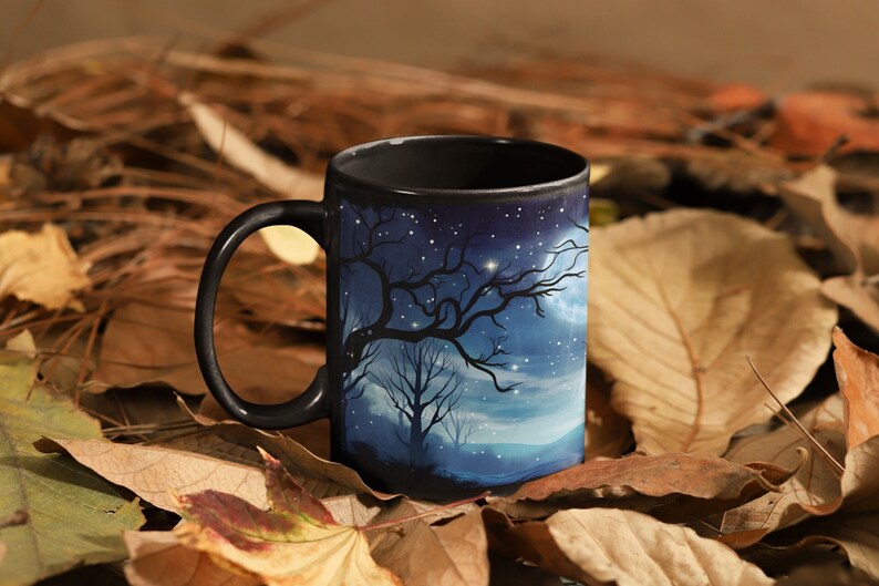 May include: A black ceramic mug with a starry night scene featuring a silhouetted tree and a crescent moon. The mug is sitting on a bed of brown and orange leaves.