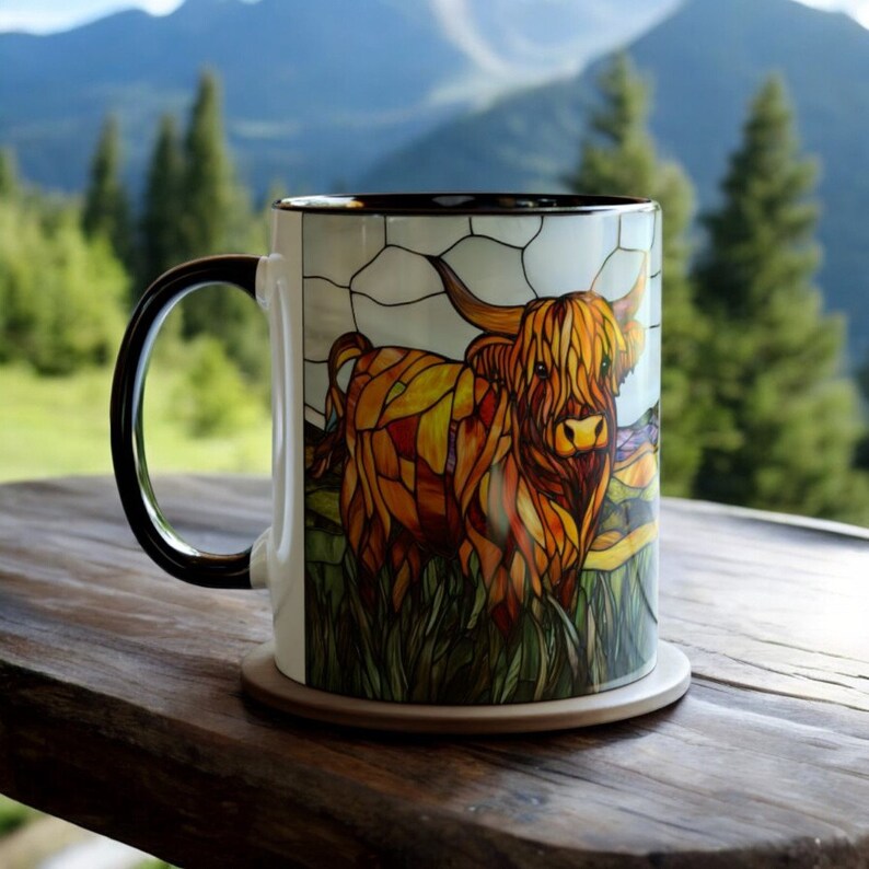 May include: A white ceramic mug with a stained glass design of a Highland cow. The cow is brown and orange with a long, shaggy mane. The mug has a black handle and is sitting on a brown wooden surface.