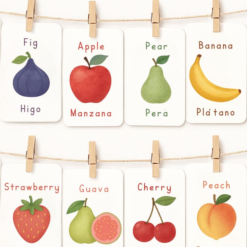 May include: A collection of illustrated fruit cards, including a fig, apple, pear, banana, strawberry, guava, cherry, and peach. Each card is labeled with the fruit's name and a corresponding word in Spanish. The cards are hung with wooden clothespins on a string.
