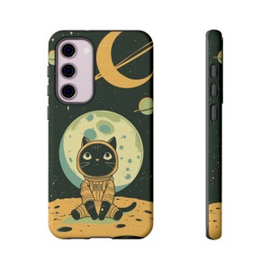 May include: A black cat wearing an astronaut suit sits on a yellow moon with a green and black space background. The phone case has a cutout for the camera lens.