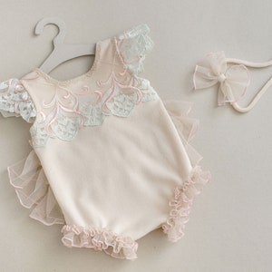 Puede incluir: Un body de bebé color crema con detalles de encaje en azul claro y rosa, colgado de una percha gris. También hay una diadema a juego con un lazo. El body presenta bordes con volantes y un diseño delicado.