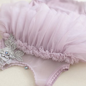 May include: A lavender-colored baby bodysuit with a tulle skirt. The bodysuit is adorned with a silver floral applique and a small, sparkling gem. The tulle skirt has a ruffled edge.