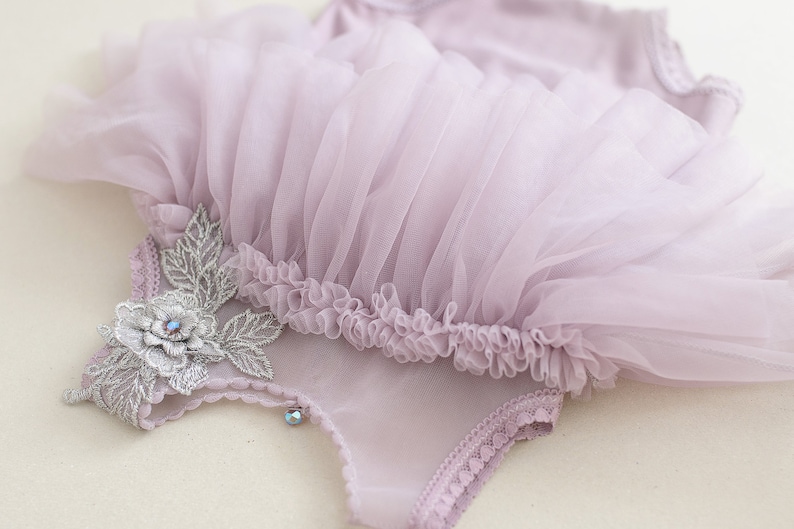 May include: A lavender-colored baby bodysuit with a tulle skirt. The bodysuit is adorned with a silver floral applique and a small, sparkling gem. The tulle skirt has a ruffled edge.