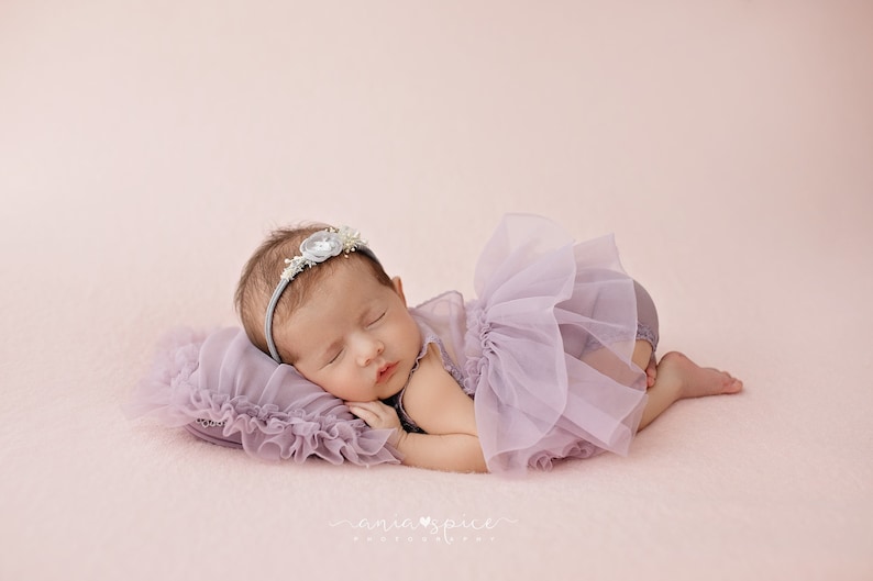 May include: A newborn baby wearing a lavender tutu and a floral headband, sleeping peacefully on a matching pillow. The baby is positioned on a soft, pink surface, creating a serene and delicate scene. The image is a studio portrait.