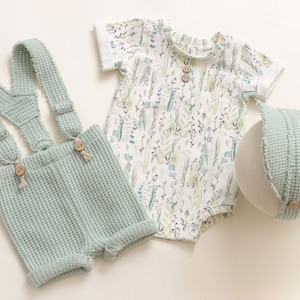 Op de afbeelding: Een baby-outfitset in lichtgroen. De set bevat een broek met bretels, een shirt met korte mouwen met een bloemenprint en een bijpassende muts.