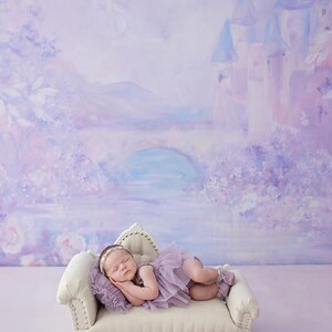 May include: A sleeping infant wearing a lavender tutu and headband rests on a small, cream-colored couch. The background features a painted mural of a castle, bridge, and flowers in shades of purple and blue. The image has a soft, dreamy aesthetic.