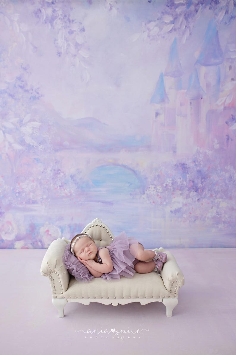 May include: A sleeping infant wearing a lavender tutu and headband rests on a small, cream-colored couch. The background features a painted mural of a castle, bridge, and flowers in shades of purple and blue. The image has a soft, dreamy aesthetic.