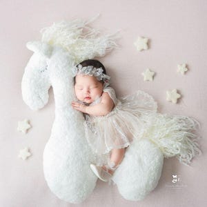 May include: A newborn baby girl wearing a white dress and a flower crown is sleeping on a white plush horse toy. The background is a light pink. There are white felt stars scattered around the horse and baby.