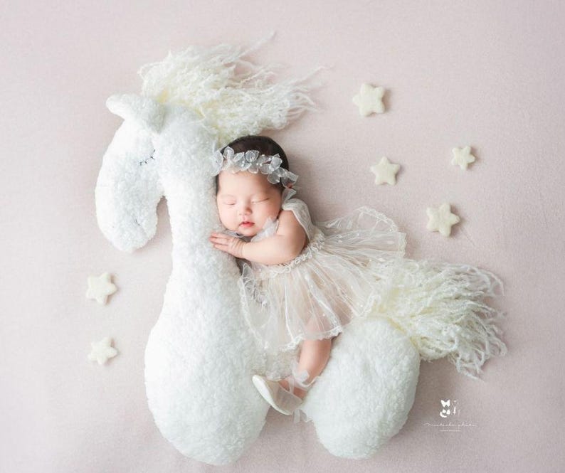 May include: A newborn baby girl wearing a white dress and a flower crown is sleeping on a white plush horse toy. The background is a light pink. There are white felt stars scattered around the horse and baby.
