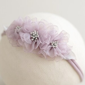 May include: A delicate headband featuring three lavender-colored fabric flowers with white beaded centers. The flowers are attached to a thin, lavender-colored band. The headband is designed for babies or young children.