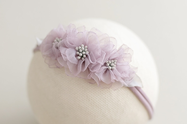 May include: A delicate headband featuring three lavender-colored fabric flowers with white beaded centers. The flowers are attached to a thin, lavender-colored band. The headband is designed for babies or young children.