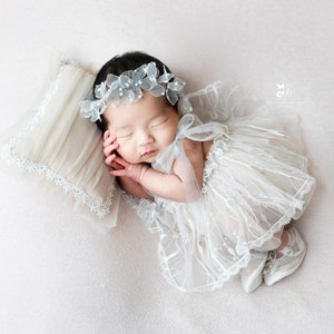 May include: A sleeping baby girl wearing a white lace dress and a flower crown. The baby is lying on a beige pillow with lace trim.