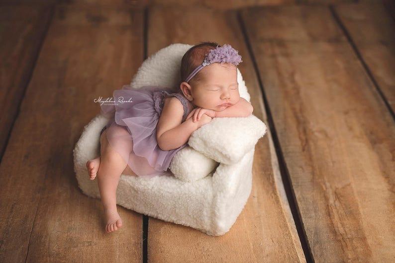 May include: A sleeping baby wearing a lavender dress and headband, resting on a small, white, fluffy chair. The background is a wooden surface with a warm, brown tone. The baby is the focal point of the image.