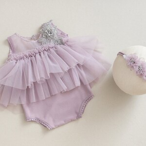 May include: A lavender baby romper with a layered tulle skirt and a matching headband. The romper features a delicate lace and floral embellishment on the shoulder. The headband is adorned with matching lavender flowers.