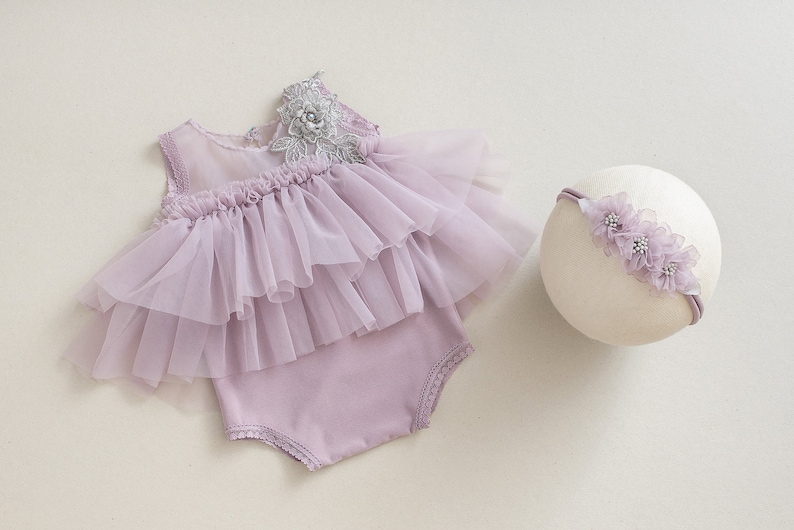 May include: A lavender baby romper with a layered tulle skirt and a matching headband. The romper features a delicate lace and floral embellishment on the shoulder. The headband is adorned with matching lavender flowers.