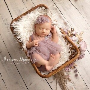 May include: A newborn baby wearing a lavender dress and headband, lying in a wooden basket lined with white fur. The basket is decorated with flowers and gold accents. The text "Precious Moments photography by MAGDA" is visible.