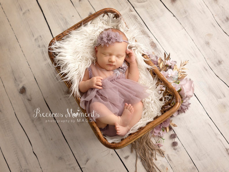 May include: A newborn baby wearing a lavender dress and headband, lying in a wooden basket lined with white fur. The basket is decorated with flowers and gold accents. The text "Precious Moments photography by MAGDA" is visible.