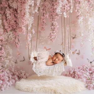 May include: A sleeping baby in a white lace swing, surrounded by pink and white flower blossoms. The swing is suspended from a white ceiling with pink and white flower garlands.