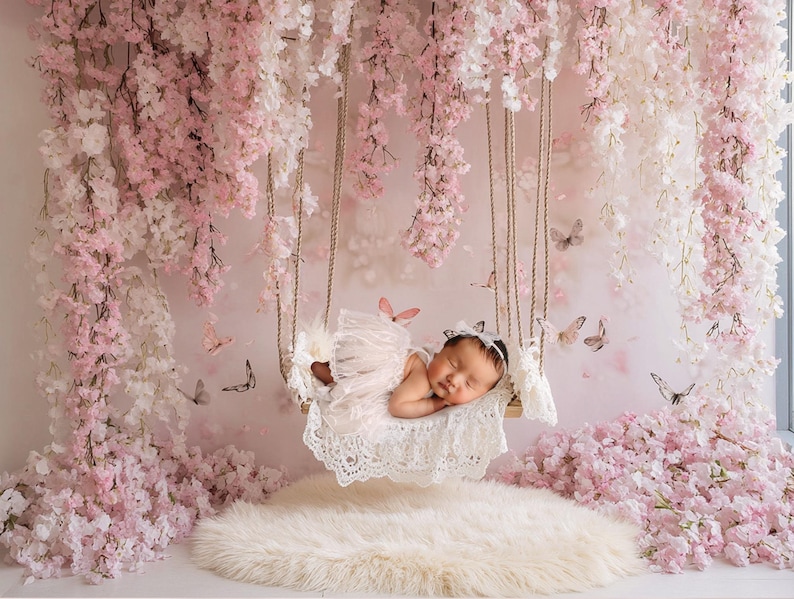 May include: A sleeping baby in a white lace swing, surrounded by pink and white flower blossoms. The swing is suspended from a white ceiling with pink and white flower garlands.