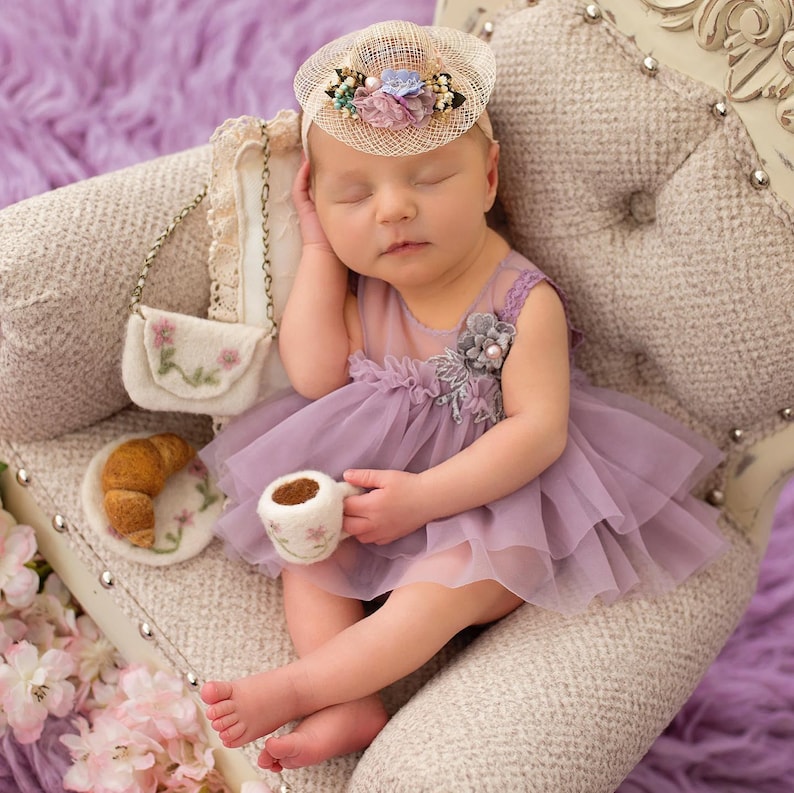May include: A sleeping baby wearing a lavender dress and a floral headband, resting on a vintage chair. A miniature purse, croissant, and teacup are placed nearby, creating a whimsical scene. The overall theme is a newborn photoshoot.