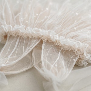 May include: A delicate white lace fabric with a ruffled edge and small sequins. The fabric is sheer and has a soft, flowing texture.